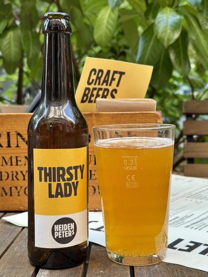 A bottle of beer labeled "THIRSTY LADY" by Heidenpeters is placed next to a clear glass filled with the same beer. The background features greenery and a sign that reads "CRAFT BEERS."