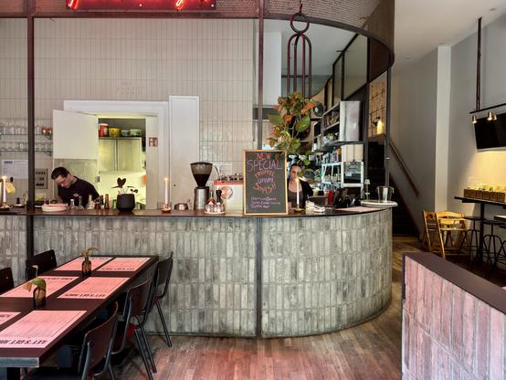 A modern cafe interior features a curved counter with two staff members, one preparing food and the other taking an order. There are potted plants and a chalkboard displaying a special menu item. The space includes wooden flooring, tables with menus, and shelves