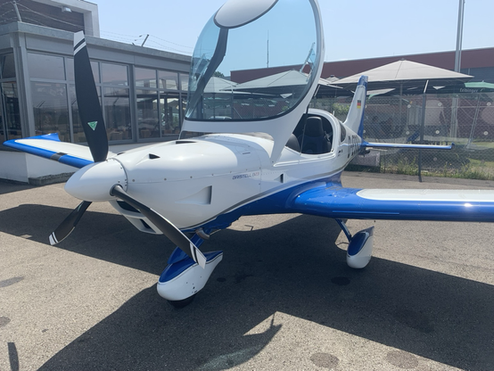 A small general aviation aircraft in white and blue standing with open canopy in front of a fence on an apron.