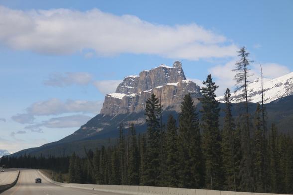 Image of a flat-topped mountain with blue sky and some wispy clouds in the background. In the foreground there is a highway and some forests.