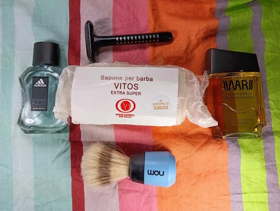 (Le Chat Mistral description)

The image displays a collection of grooming items arranged on a striped, multicoloured fabric. At the top left, there is a bottle of Adidas Dynamic Pulse Eau de Toilette. Next to it, towards the top centre, is a black razor. In the middle, there is a wrapped bar of soap labelled "Vitos Extra Super" with some text in Italian. To the right of the soap, there is a bottle of Wars Classic aftershave lotion. At the bottom centre, there is a shaving brush with a blue and black handle that has the word "wou" written on it. The items are neatly laid out, suggesting a shaving or grooming kit.
