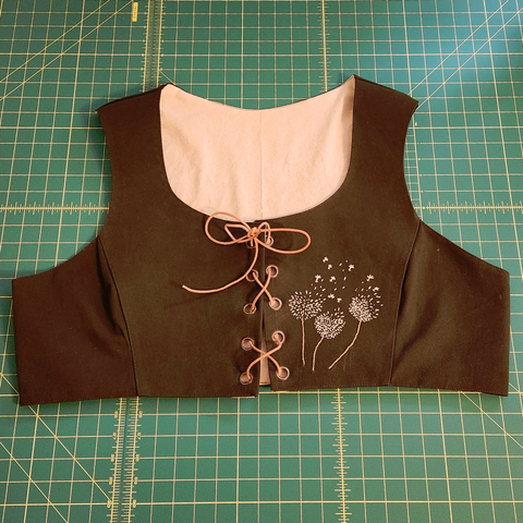 An olive green cropped bodice vest that is laced up with a leather cord. On the one side of the front are three embroidered dandelion puffs with some of the seeds scattered up towards the shoulder.