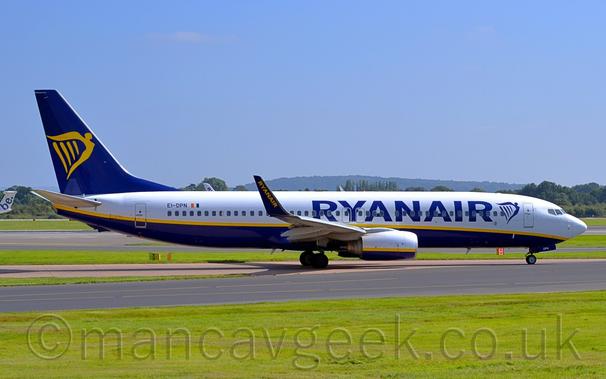 Side view of a twin engined jet airliner taxiing from left to right along a grey taxiway.
The plane is mostly white on top, with a dark blue belly and a yellow stripe swooping from under the nose to the upper rear fuselage, repeated on the engine pods mounted under the wings.
There are large, dark blue "RyanAir" titles on the forward fuselage, next to a blue, winged Irish harp.
The tail is dark blue, with a large, yellow, winged Irish harp.
The up-turned wingtips are the same dark blue, with diagonal yellow "RyanAir" titles.
Vibrant green grass lines the sides of the taxiway in the foreground, with trees and vivid blue sky in the background.