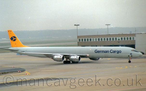 Side view of a long, thin, 4 engined jet airliner taxiing from left to right.
The plane is mostly a dark grey, with a lighter grey belly, and dark blue "German Cargo" titles on the forward fuselage.
The tail is bright yellow, with a logo of a 3-D blue box in the middle - the same logo appears in a smaller form under the cockpit windows.
Grey concrete apron fills the foreground, with a brown building stretching half way across the frame from the right in the background.
On the left, grass and runways lead up to trees in the distance, under hazy grey skies.