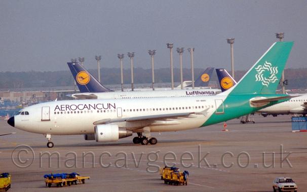Side view of a twin engined jet airliner taxiing from right to left.
The plane is mostly white, with black "AeroCancun" titles on the upper forward fuselage.
The rear fuselage are a minty green, with a white stylised flower logo made up of six sets of 3 wavy lines arranged in a circle.
Grey concrete apron fills the foreground, with yellow luggage carts with blue covers parked at the side of a runway marked on the concrete.
In the background, several white jet airliners with blue and yellow tails are parked amidst some tall lighting towers, with trees in the distance, under grey sky.