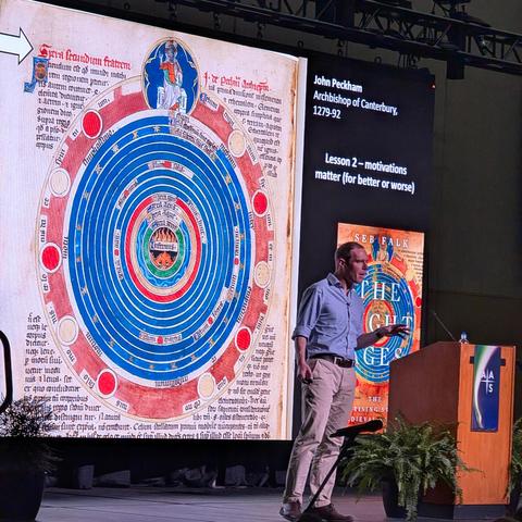 Seb Falk stands on stage. Behind him is a giant screen with a medieval illustration of the spheres of the heavens, with the Earth at the center, and a series of concentric rings for the Sun, Moon, Earth, planets, and the fixed stars.
