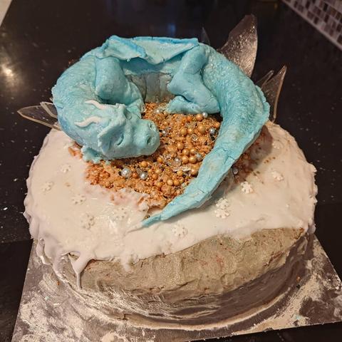 A cake topped with an ice blue dragon, sleeping on a little pile of treasure. The cake itself is a snowy rock. Behind the dragon are shards of ice, sticking upwards.