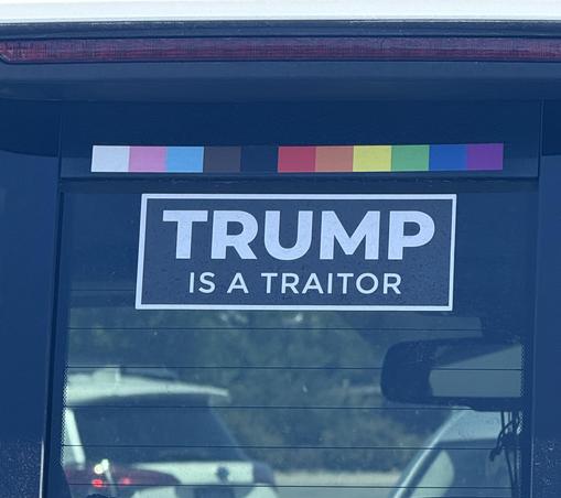 A window decal on a car says in all capital letters: TRUMP IS A TRAITOR.  There's a bar of rainbow colors on top.