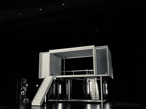 Dunkle Theaterbühne vor Beginn des Stücks, darauf eine zweigeschossige Installation auf Stützen, die obere Ebene stellt ein offenes Zimmer mit weissen Wänden dar, eine ebenfalls weiße Treppe führt von vorn hinauf.