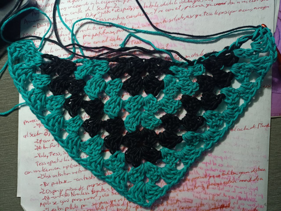 Chal con otro vueltas en el que voy alternando negro con verde azulado. La pieza crece en forma triangular y las franjas tienen forma de V.
Por encima le sobresalen un montón de hilos de ambos colores.
Está colocado sobre unos folios escritos en rojo.