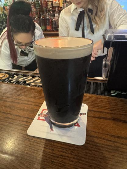 A dark beer is placed on a coaster at a bar. In the background, two bartenders are working, one with long hair is leaning over, while the other is preparing drinks. Shelves filled with various bottles are visible behind them.