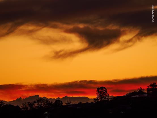 A dramatic and intense dawn sky. There are three cloud layers seen: just visible behind the dark suburban horizon is a distant low bank of dark grey cumulus; immediately above is a line of flat cloud, lit an intense red by the unrisen sun; above that again is a clear, orange-yellow sky, and then capping the image is a dark, wispy, unlit bank of cloud scudding across the sky