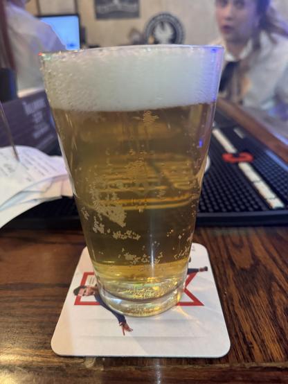 A glass of beer with a frothy head rests on a coaster featuring an image of a person, on a wooden bar surface. The background includes blurred elements of a bar setting.