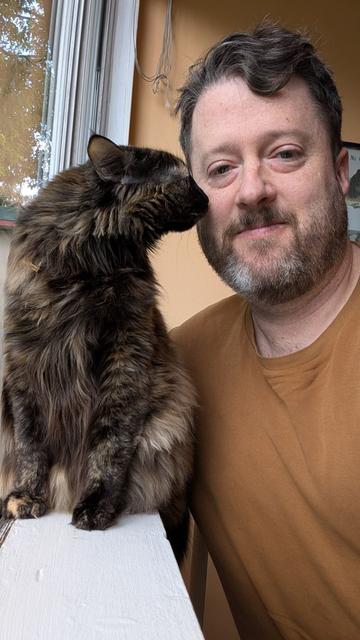 Selfie of a guy with a longhaired tortoiseshell cat. The cat is leaning over to smell his cheek