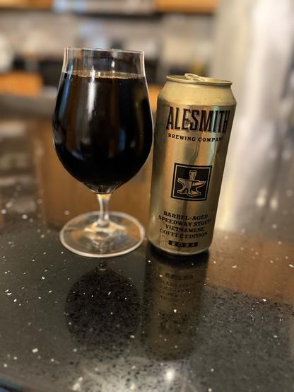 A large snifter glass chock full of dark beer. The golden pint can is to the right with the gold-on-black AleSmith anvil logo