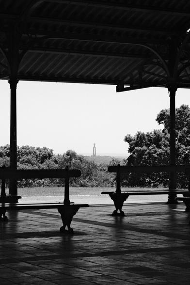 BW photo of the Cristo Rei statue, as seen from the Miradouro Alto da Serafina, in Monsanto. In the foreground a few empty benches.