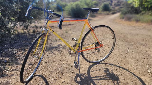 Bright yellow and orange painted bike with blue brake and shift cables. Brand new tires, everything is shiny. Photo taken in late afternoon, backlight, a hilly area/trail behind with trees and bushes. Shadow is long and comes towards the photographer.