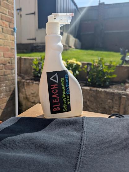 A white spray bottle with a hand written black label on it, on a table in a sunny garden. The label reads "Bleach. Sodium Hypochlorite 4.5%. 50% diluted."