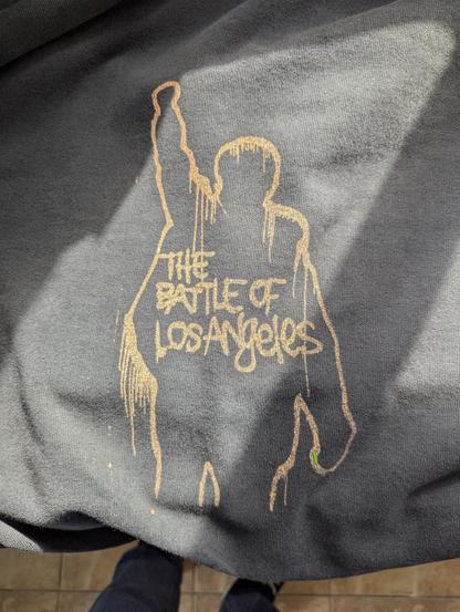 A close up of the back of the hoody, showing the finished outcome. An outline of a person with a raised fist in the air, the outline appears to be dripping with paint. Inside the outline the words "The Battle of Los Angeles" in all caps graffiti style, as if written with a can of spray paint