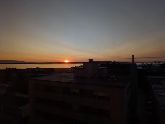 Sunset on a clear day. The city is in shadow in the foreground. In the background is the Cook Inlet and distant mountains.