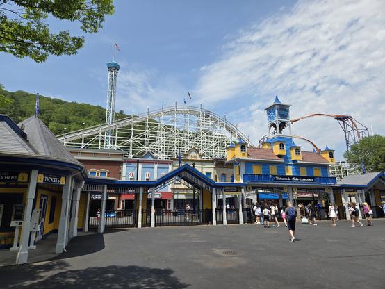 The front entrance gate of Lake Compounce amusement park, a festive yellow-and-blue structure with turnstiles beneath. Behind it and some other brightly colored buildings rises the white-painted lift hill of a wooden roller coaster; behind that, an orange steel coaster and a tall drop tower; behind those, a forested mountainside.