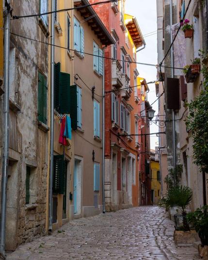 A quiet, narrow cobblestone street in Rovinj, Croatia, glows with soft morning light, flanked by colorful pastel buildings with shutters, hanging laundry, and flower pots that hint at daily life in this historic coastal town.