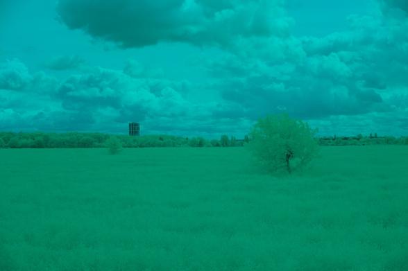 The image features a landscape with a grassy field under a cloudy sky. In the distance, there is a tall structure, possibly a tower, rising above a line of trees. The overall color tone of the image is greenish-blue, giving it a surreal appearance.