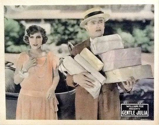 There is a man and a woman. The woman is wearing a short-sleeved pink dress and a necklace. Her hair is short, dark and wavy. The man is holding lots of packages. He is wearing a brown suit and a white hat with a black stripe.