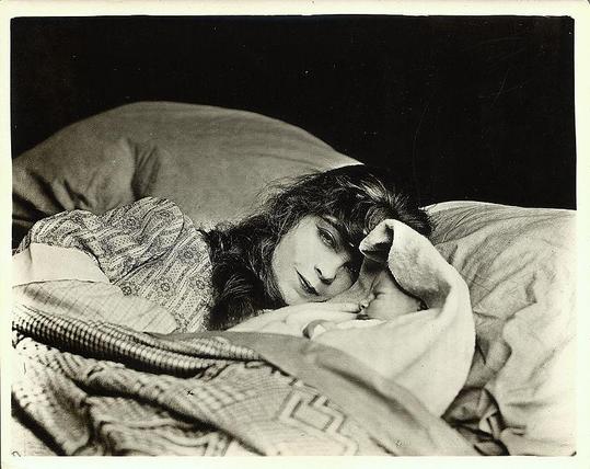 A woman is laying down a bed besides a baby. She has long, dark wavy hair. There are two white pillows and they are under gray blankets.
