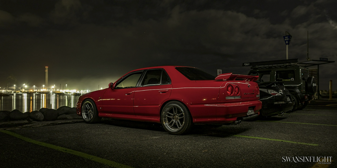 A red R34 Nissan Skyline lit against a night sky backdrop of the New Plymouth port, cranes and the like lit up in the background beyond the water between.