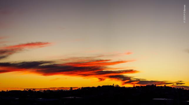 A train of blown, wispy cloud trails across the sky like blown smoke. It's lit an intense red-orange by the unrisen sun. The land is a dark silhouette, as the sky behind brightens to yellow below and slate blue above.