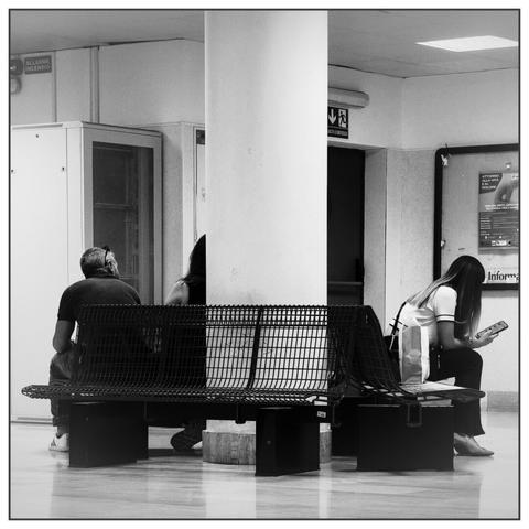 Fotografia in bianco e nero di una sala d’attesa. In primo piano una panchina in metallo scuro, su cui sono sedute tre persone: due sul lato sinistro, separate da una colonna, e una ragazza sulla destra intenta a guardare il telefono. L’ambiente  ospedaliero, con pareti chiare, pavimento lucido e segnaletica visibile.