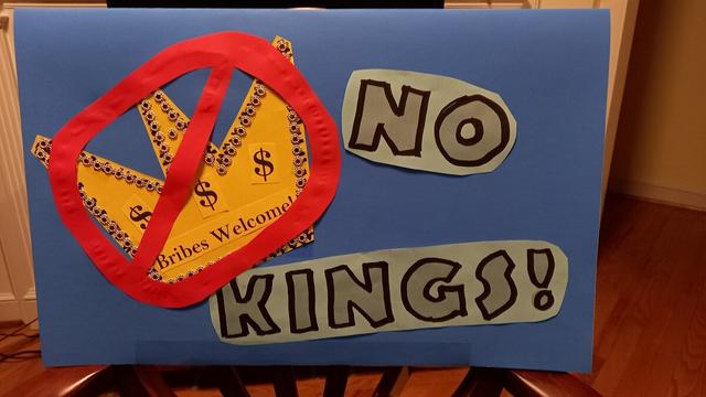 Protest sign -- on a piece of blue posterboard, landscape format, my take on the No Kings crown logo with a red circle-slash over it. The crown is on gold paper with sequins, and inside it has dollar signs and "Bribes Welcome" -- but it's got the "no" slash over it. To the right of that is the phrase NO KINGS! in block letters, cut out of light blue paper and outlined in black. It's very arts-and-craftsy, lo-tech paste-up