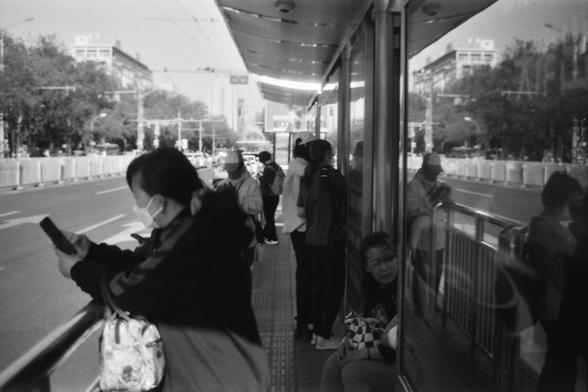 Ilford FP4 E.I. 64

**English:**
This black-and-white photograph captures a scene at a bus stop. Several people are waiting, some standing and some sitting. A woman in the foreground is engrossed in her smartphone, wearing a mask and carrying a bag. The bus stop shelter is visible, and the street outside is lined with trees and buildings, indicating an urban setting. The overall atmosphere suggests a typical day in a city with people going about their daily routines.

**Chinese:**
这张黑白照片捕捉了一个公交车站的场景。几个人在等车，有些站着，有些坐着。前景中的一位女性正专注于她的智能手机，戴着口罩并提着一个包。公交车站的候车亭清晰可见，外面的街道两旁种满了树木和建筑，显示出一个城市环境。整体氛围表明这是城市中人们日常生活的一个普通日子。