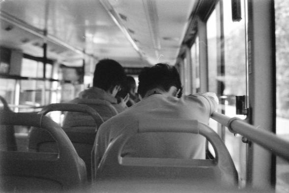 Ilford FP4 E.I. 64

**English:**
This black-and-white image shows the interior of a bus. The focus is on the backs of two passengers seated near the front. The bus is in motion, as suggested by the blurred scenery visible through the windows. The interior lighting is bright, illuminating the passengers and the details of the bus seats and handrails. The image conveys a sense of movement and daily commuting.

**Chinese:**
这张黑白图片展示了公交车内部的场景。焦点集中在靠近前部的两名乘客的背影上。公交车正在行驶中，从窗外模糊的景色可以看出这一点。车内的灯光明亮，照亮了乘客以及公交车座位和扶手的细节。这张图片传达了一种运动感和日常通勤的氛围。