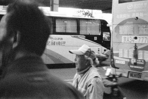 Ilford FP4 E.I. 64

**English:**
This black-and-white image captures a moment at a bus station. In the foreground, a woman wearing a cap is seen from the side, possibly talking or looking at something off-camera. Behind him, a large bus is parked, with visible text on its side. The bus station appears busy, with other people and vehicles in the background. The scene suggests a bustling transportation hub with people in transit.

**Chinese:**
这张黑白图片捕捉了公交车站的一个瞬间。前景中，一个戴帽子的女人从侧面可见，可能正在说话或看着镜头外的某物。他身后停着一辆大巴，侧面有可见的文字。公交车站看起来很繁忙，背景中还有其他人和车辆。这个场景表明这是一个繁忙的交通枢纽，人们正在途中。