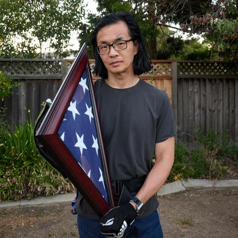 Me, an Asian guy with shoulder length long hair and square glasses holding a triangular frame showing a folded flag in the tricorn shape from military funerals.