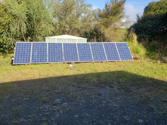A row of 8 solar panels on the edge of a grassy yard. Behind is a small shed and lots of bush
