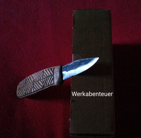 Ein Messer ist so ins Licht gesetzt, dass die Schneide glänzt. Es steht angelehnt an eine Pappkiste auf einem roten Tuch und wurde von oben fotografiert.