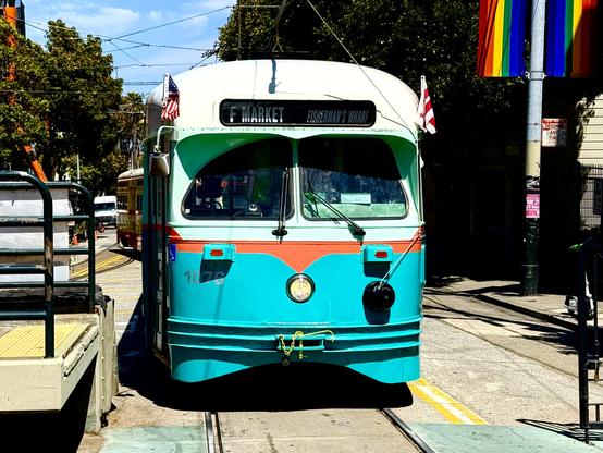 A teal and cream-colored historic streetcar labeled “F Market” with a destination sign reading “Fisherman’s Wharf” is parked on streetcar tracks in San Francisco. The car has an American flag and another red-striped flag mounted on each side. A rainbow pride flag hangs from a pole on the right, and another vintage streetcar is visible further down the tracks in the background. Trees and urban infrastructure frame the scene under a sunny blue sky.