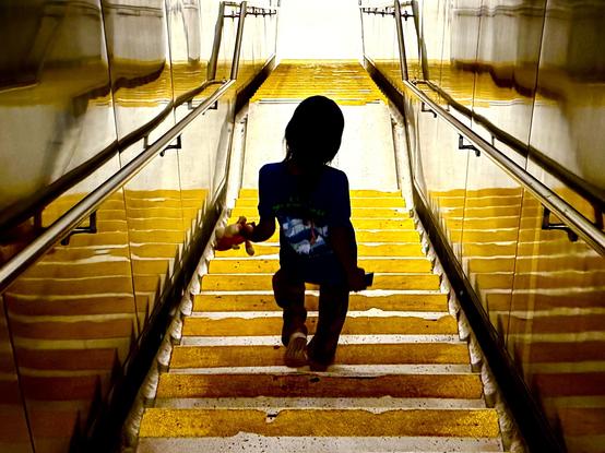 A child is descending a stairway that is painted in alternating yellow and white stripes. The child is in silhouette, holding a toy in one hand. The stairwell is flanked by metallic walls that reflect the yellow stripes, creating a distorted mirror effect. The lighting is bright at the top of the stairs, casting the lower area into deeper shadow.