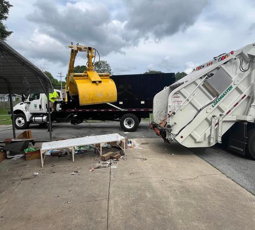 a photo outside of the cities sanitation crew's trucks. there is someone operating the claw. which is positioned above a table, and many scattered & broken objects all around on the concrete floor.