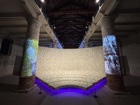 Frontal view of the large curved brick wall between columns with projected images. The bricks form a steep slope. Blue and purple light illuminates the space.