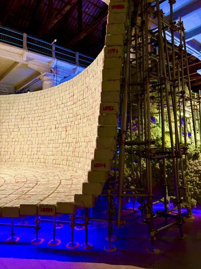 Large curved wall made of pale, brick-like blocks supported by metal scaffolding. The wall is marked with red years (1651, 1804, 1927, 1974, 2025), showing population or historical milestones. Purple light glows beneath.