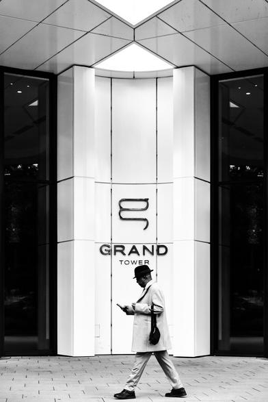 black and white. portrait format. an elegantly dressed gentleman with a hat walks past the entrance area of a modern residential tower. On the facade is the name of the building: Grand Tower. And in front of him the Grand Seigneur