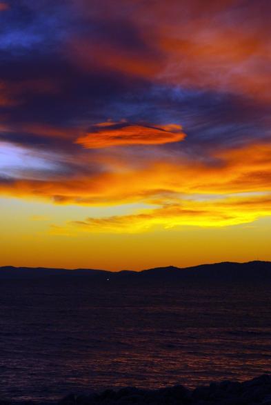 Photo au format vertical d’une plage juste après que le soleil a disparu derrière l’horizon. La mer est d’un beau violet sombre avec des reflets rose-orangés, et est bordée dans le fond par des collines basses qui se découpent en noir devant le ciel jaune orangé. Au dessus, des nuages comme peints par la lumière, avec un dégradé de rose, violet sombre, orange et jaune.