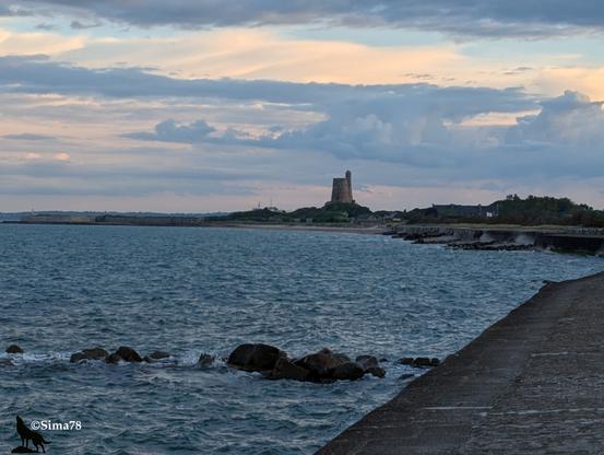 Sous un ciel nuageux de gris foncé à blanc avec quelques percées bleues.
La mer. Le quai file en bas sur la droite pour revenir en face au centre de la photo où se trouve le fort de la Hougue,