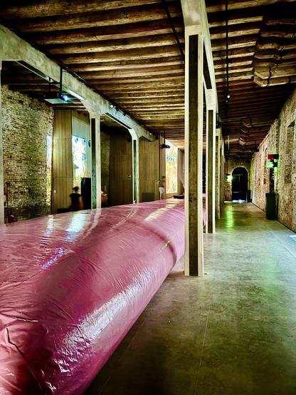 Long, narrow exhibition hall with brick walls and wooden-beamed ceiling. In the center stretches a massive, shiny pink inflated structure, like a soft, air-filled tube, running almost the full length of the space. The polished floor reflects subtle colors from projections on the walls. The atmosphere blends industrial rawness with surreal softness.