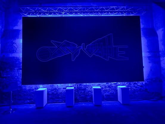 Dark exhibition space bathed in intense blue light. A large black panel displays the word Oxyville in geometric, futuristic typography, resembling interconnected architectural forms. Four white plinths and a single chair stand below the panel