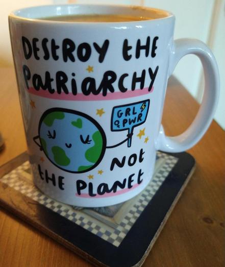 Mug of tea on a bedside cabinet, the slogan 'Destroy the Patriarchy ' at the top cartoon globe in the middle and the words 'not the planet ' at the bottom